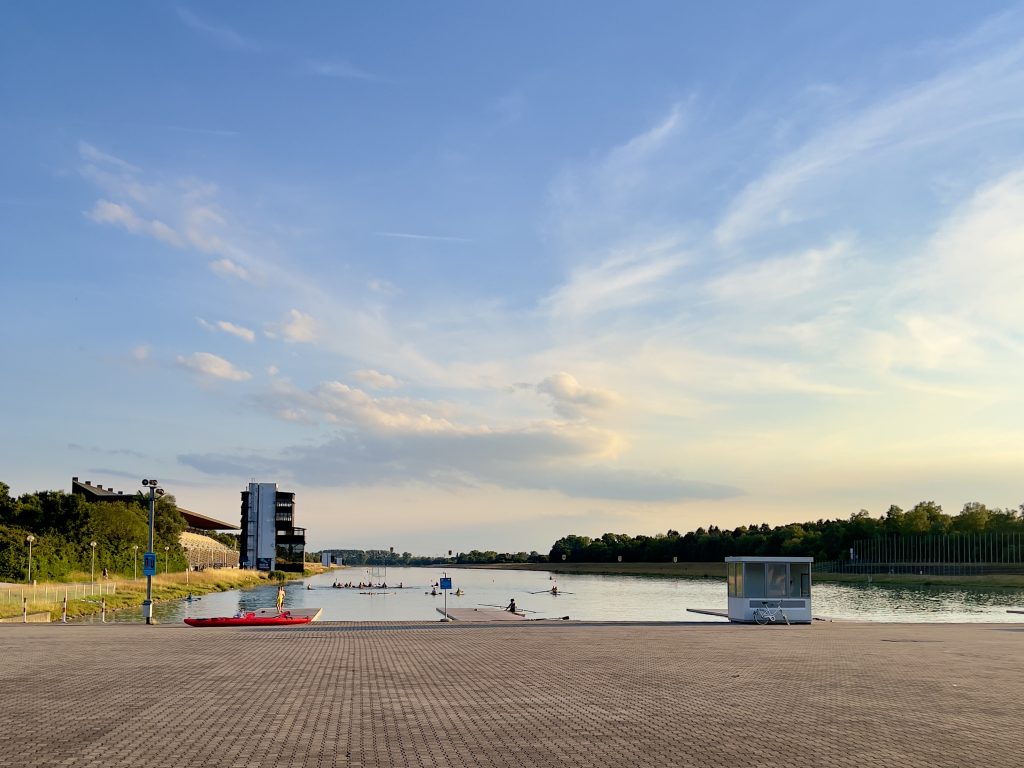 Die Regattastrecke in Oberschleißheim mit einer Zuschauertribüne am linken Bildrand. Im Hintergrund sind Ruderer auf dem Wasser aktiv.